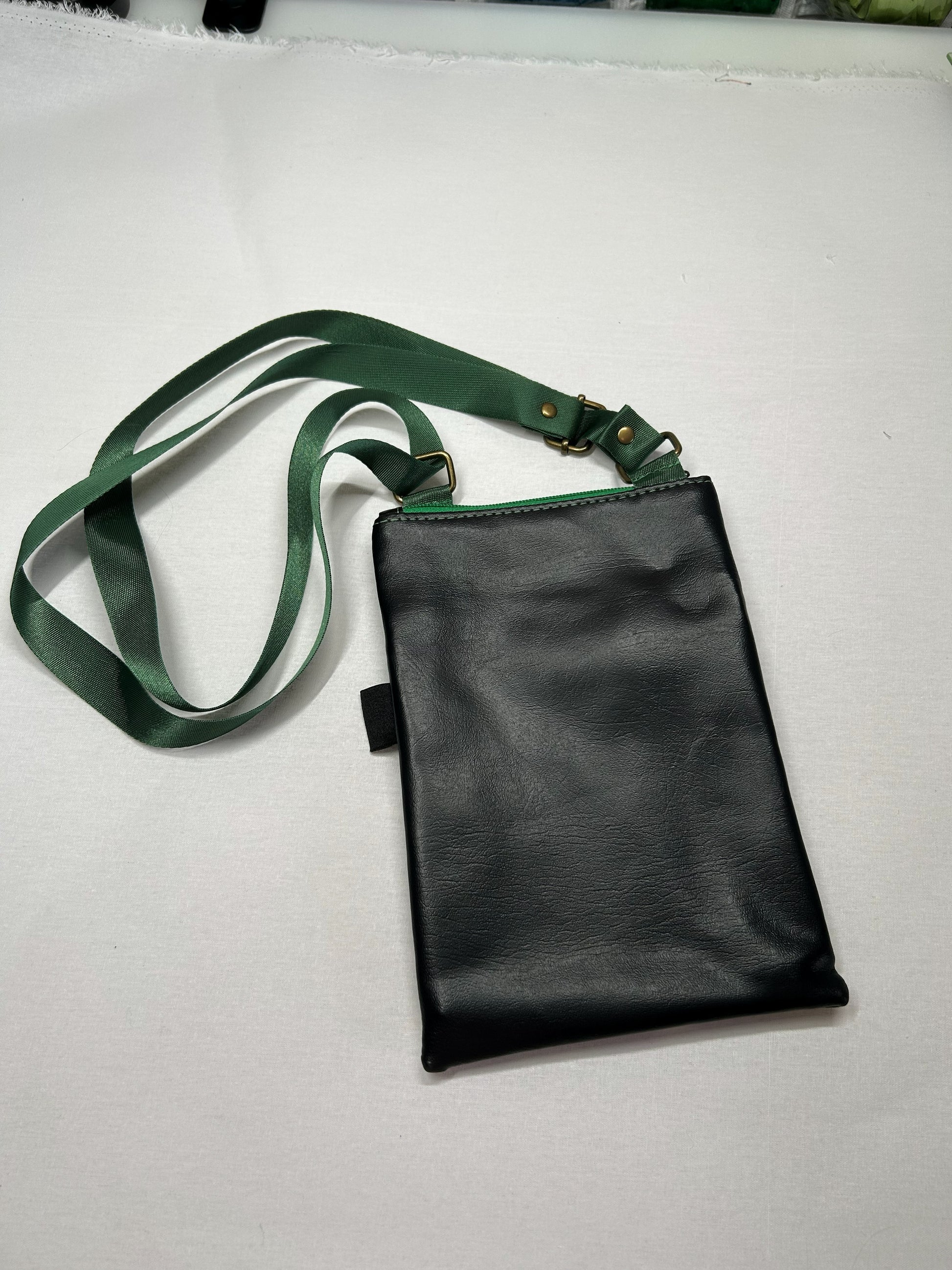 A black leather crossbody bag with a green strap is shown on a white surface.